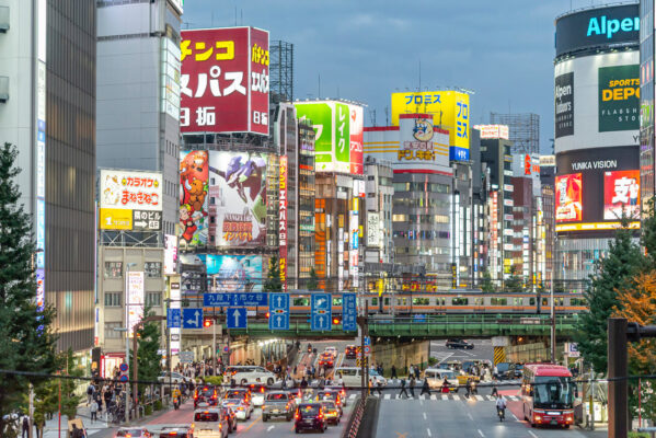 Shinjuku Tokyo cityscape at dusk with neon billboards, city traffic and a passing train above the junction seen in the opening of Midnight Diner Tokyo Stories.