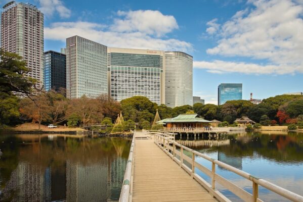 Wooden bridge leading to Nakajima Tea House across the pond at Hamarikyu Gardens in Tokyo, with Japanese garden trees and modern skyscrapers in the background.