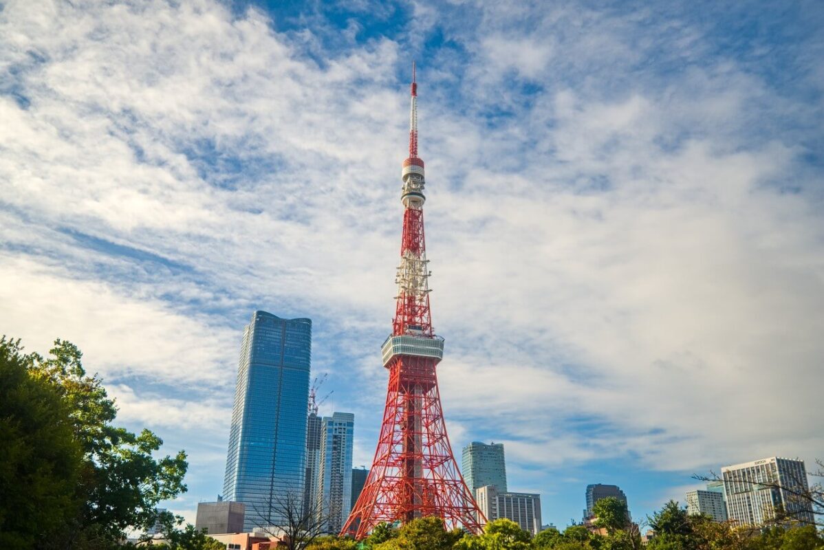 Tokyo Tower from Shiba Park in Tokyo, Japan, rising above trees and nearby high-rises under a cloudy blue sky.