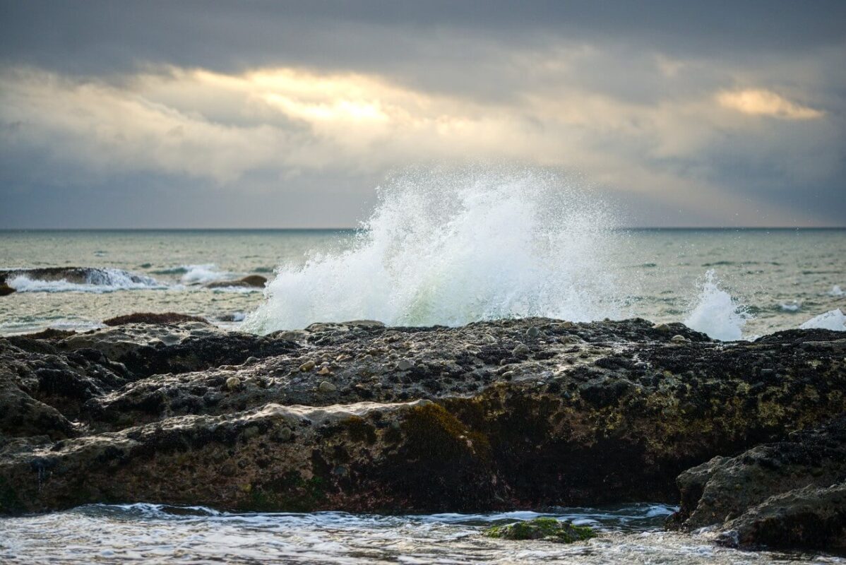 Ocean wave crashing over rocks at sunrise under a cloudy sky in Oarai Park, Ibaraki, Japan.