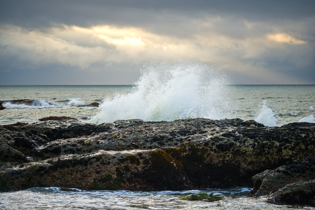 Ocean wave crashing over rocks at sunrise under a cloudy sky in Oarai Park, Ibaraki, Japan.