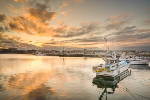 Fishing boats at sunset in Nakaminato Port, Ibaraki, Japan, with a white boat in the foreground, waterfront buildings and orange clouds reflected across the calm harbor.