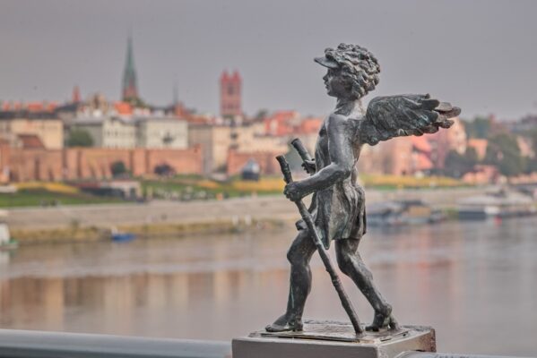 Neucjusz, a small bronze angel holding walking poles on a bridge, with the Vistula River and Torun's Old Town softly blurred in the background.