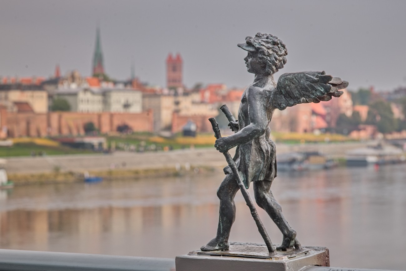 Neucjusz, a small bronze angel holding walking poles on a bridge, with the Vistula River and Toruń's Old Town softly blurred in the background.