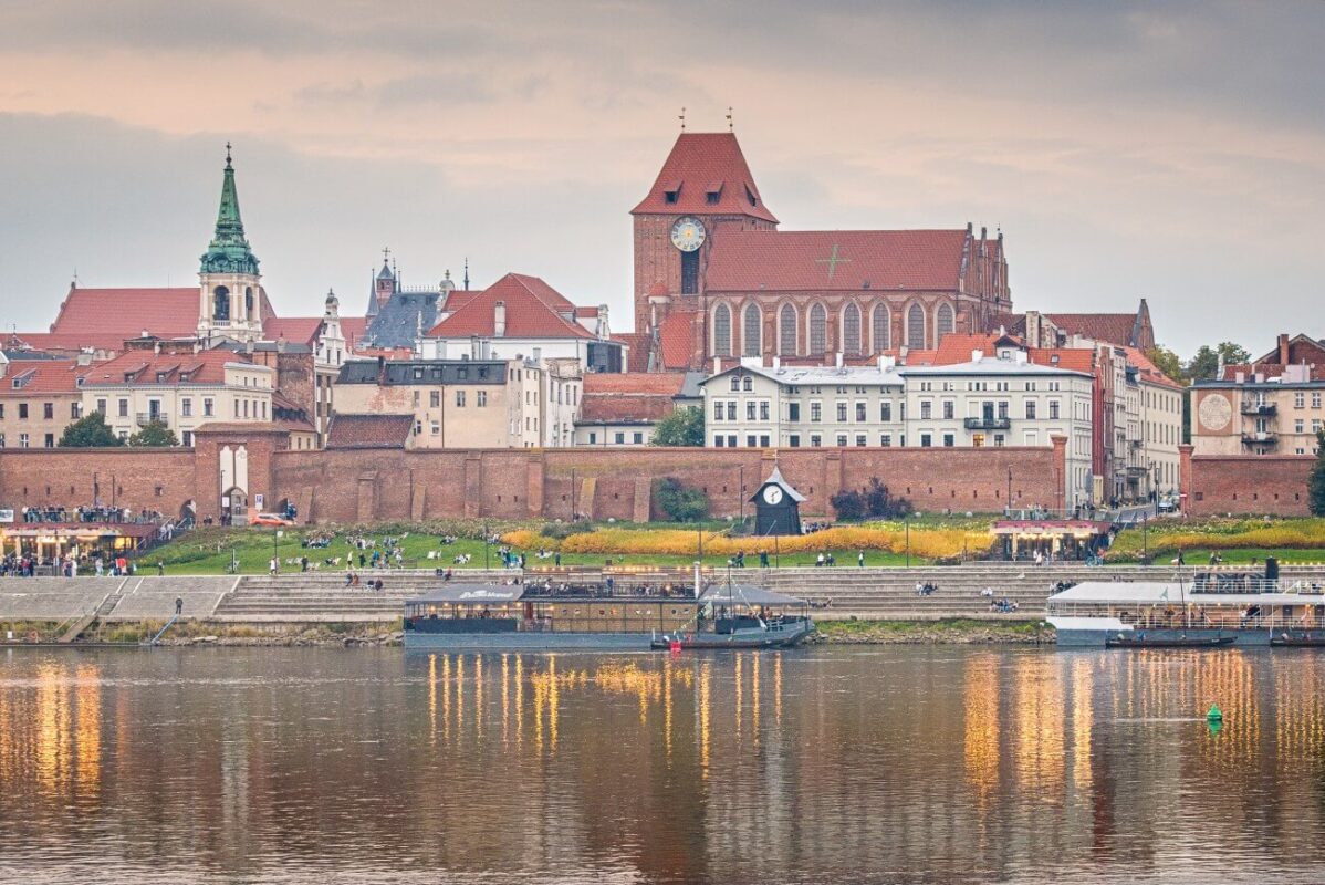 Torun Old Town at sunset across the Vistula River, with Gothic church towers, medieval walls and warm reflections on the water.